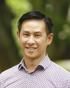 A headshot of Eugene Wong with a blurred green background. Eugene is a man of Asian appearance, side-parted short hair and is wearing a button up shirt.