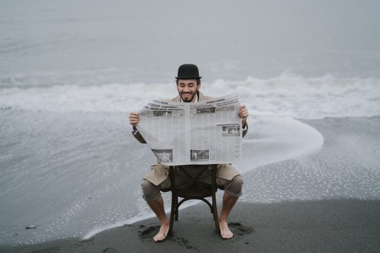 A man wearing a top hat sits on a chair at the beach, reading a newspaper
