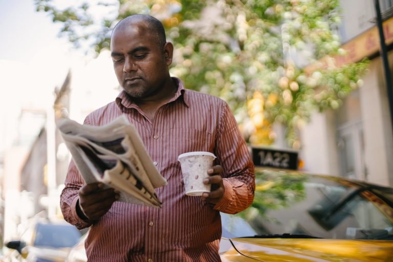 A man walks outside while reading a newspaper and holding a takeaway coffee.