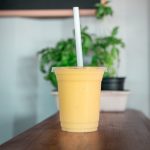 A mango lassi in a take away container with a straw sits on a bench.